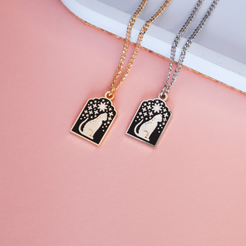 Two cat necklaces, one gold and one silver. The designs are doorway shaped and feature a sitting cat. Raining down on the cat are lots of stars with a large star centrally placed. It features black and white enamel.