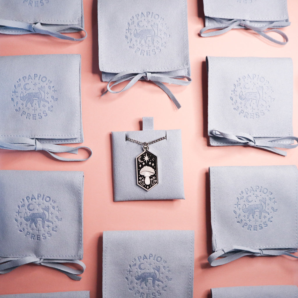 A necklace on a fabric backing. Surrounded by blue embossed Papio Press fabric pouches.