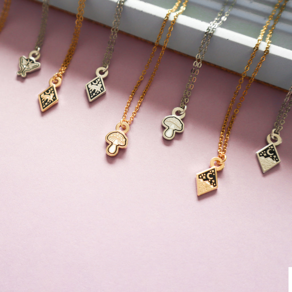 A collection of mini necklaces including the fox, mushroom, moth and mountains designs all on a blush pink background.