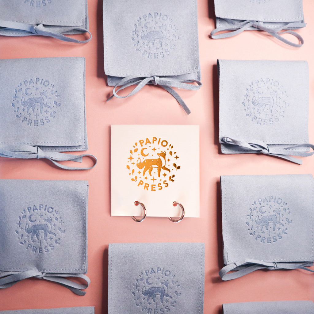 A pair of silver huggie hoops on a white Papio Press backing board surrounded by blue fabric pouches.