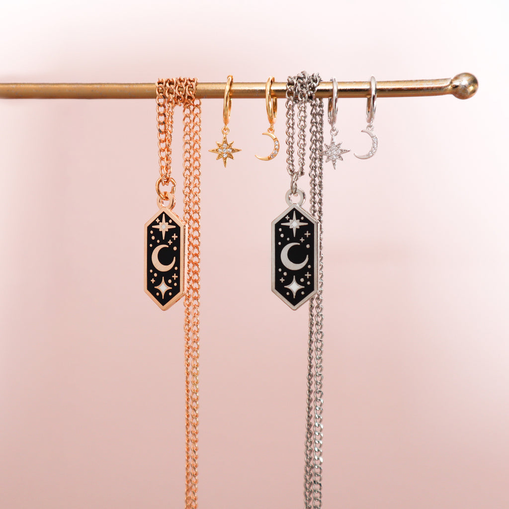 Two pairs of Celestial Moon and Stars earrings paired with the Crescent Moon necklace on a pink background.