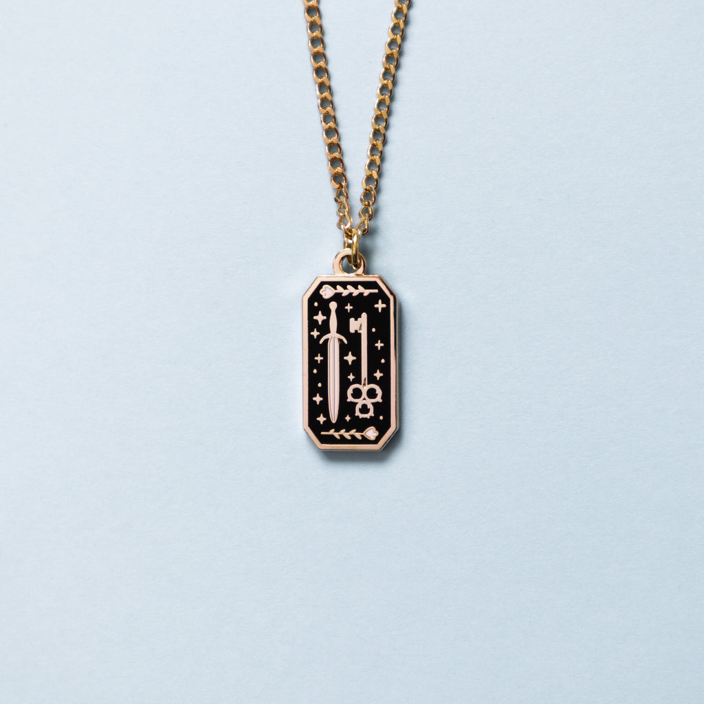 A gold rogue necklace, the necklace is rectangular shaped with a dagger and key. Above the dagger and key are flowers with stars dotted throughout the design.