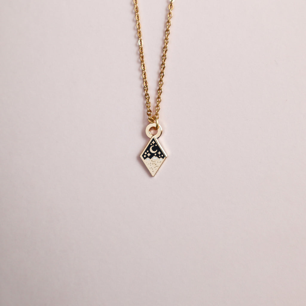 A gold mini mountains necklace on a blush pink background.