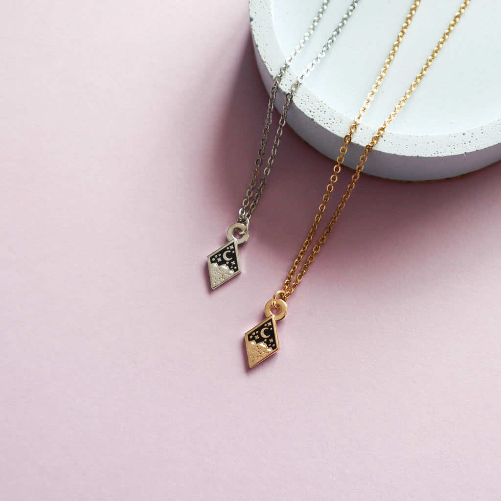 Two mini mountains necklaces, one gold and one silver. The designs are diamond shaped and feature some mountains, a moon and stars with black and white enamel details.