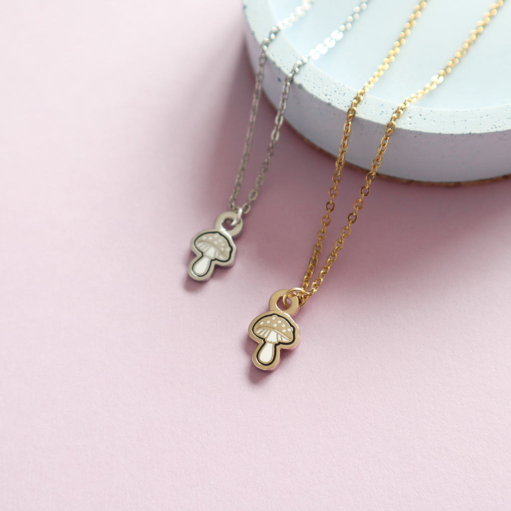 Two mini mushroom necklaces, one gold and one silver. The designs are mushroom shaped and feature black and white enamel details.
